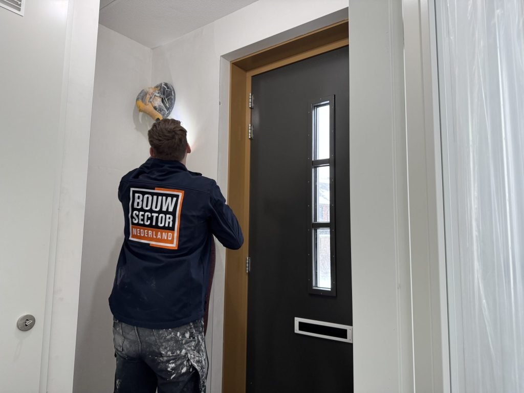 Vakman van Bouwsector Nederland schuurt met een schuurmachine op een steel de bovenkant van een wand in een hal naast de voordeur