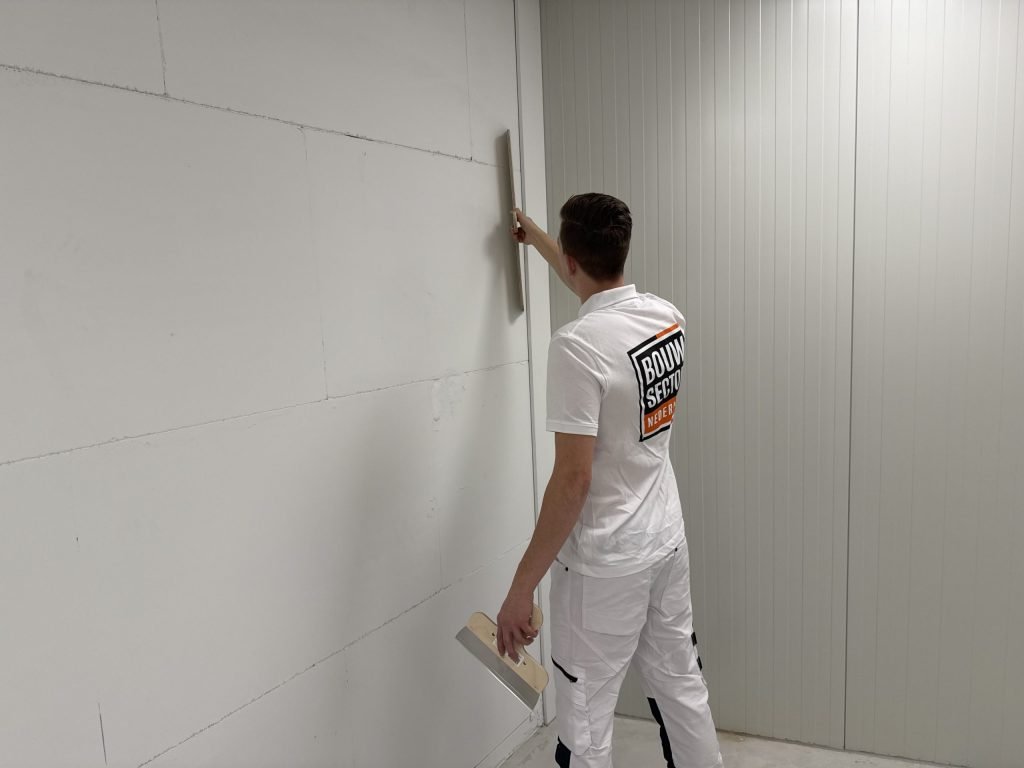Stukadoor houdt een lange spaan verticaal tegen een wand van gipsblokken, met een brede plamuurspaan in zijn andere hand