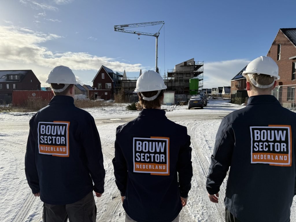 Drie vakmensen van Bouwsector Nederland met witte helmen op een besneeuwde bouwplaats met nieuwe woningen en een bouwkraan