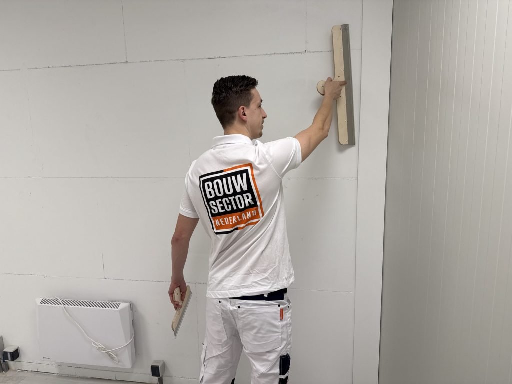 Stukadoor van Bouwsector Nederland trekt met een lange spaan de bovenkant van een wand vlak, met een kachel ernaast